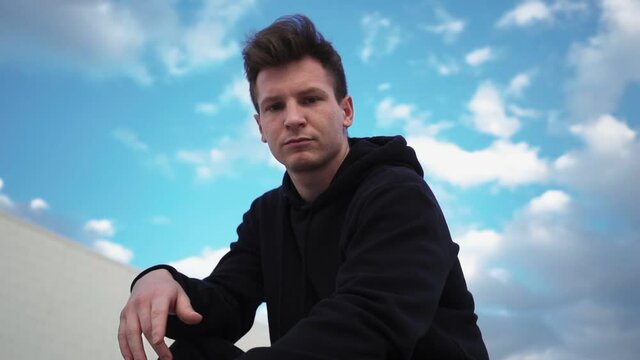 Stylish man in a black hoodie sits in a city park and looks at the camera. Camera Rotates 360 Degrees. Slow motion. 