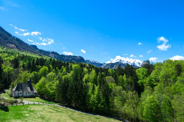 Fototapeta na wymiar A a small chapel at the edge of the forest the panoramic view on Baeren Valley in Austrian Alps. The highest peaks are snow-capped. Lush green pasture. Clear and sunny day. High mountain chains.