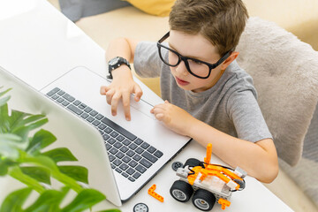 Clever young schoolboy in glasses builds and programs a robotic vehicle codes an electronic toy at home. Child using laptop.