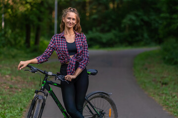 Portrait of a smiling young woman in a shirt and leggings on a bicycle standing on an asphalt road in a park on a summer day and looking at the camera, copy space