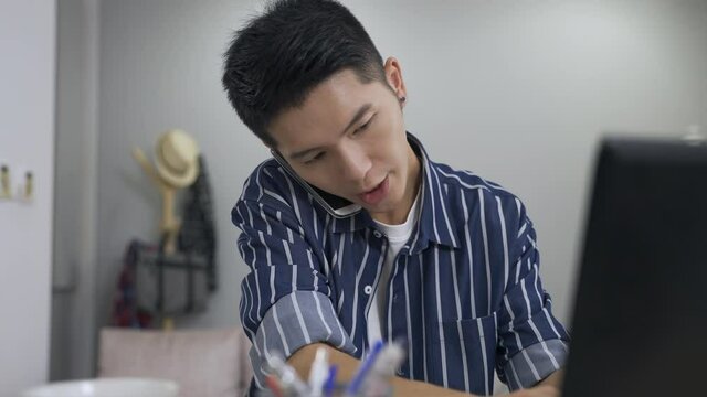 asian creative worker holding phone between ear and shoulder is writing notes and looking at the computer while having a discussion with his colleague at home.