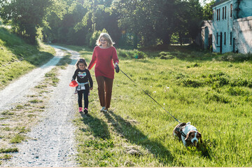 Trendy senior lady and her little toddler girl enjoying a walk in the countryside with their beagle...