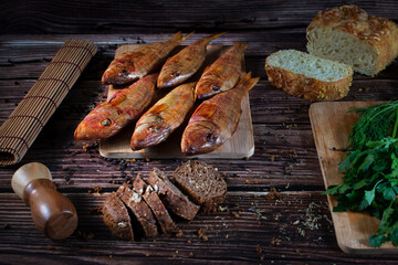Smoked red mullet fish is laid out on a wooden board, next to white and black bread, fresh herbs. Side view, brown background.