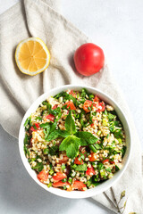Oriental salad Tabouleh of boiled bulgur, served in a white plate on a linen napkin, next to a tomato and a slice of lemon. On a white background, close-up, top view.