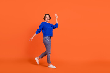 Full length body size view of attractive cheerful girl walking showing v-sign isolated over bright orange color background