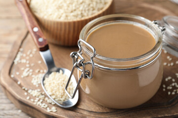 Jar of tasty sesame paste and seeds on wooden board, closeup