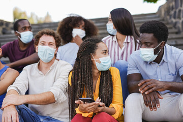 Group of young multiracial friends sitting on stairs in the city while wearing surgical face mask for coronavirus outbreak - Mixed race people enjoy time together outdoor
