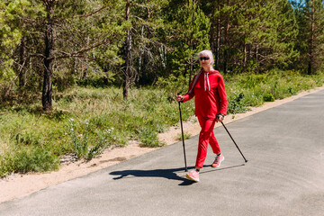 an elderly woman in red tracksuit walks with Nordic walking sticks in the summer