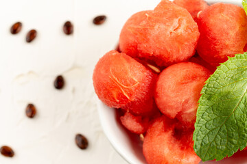 Top view of white bowl with watermelon balls and mint leaf, on white table with seeds, horizontal