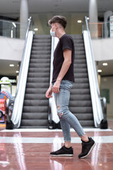 Fototapeta na wymiar Chico joven caminando en el centro comercial con mascarilla con las escaleras mecanicas