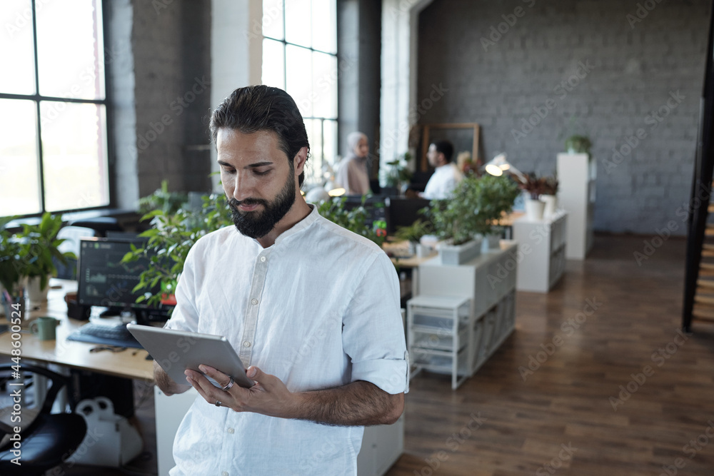 Poster young serious businessman with tablet scrolling through data in large office