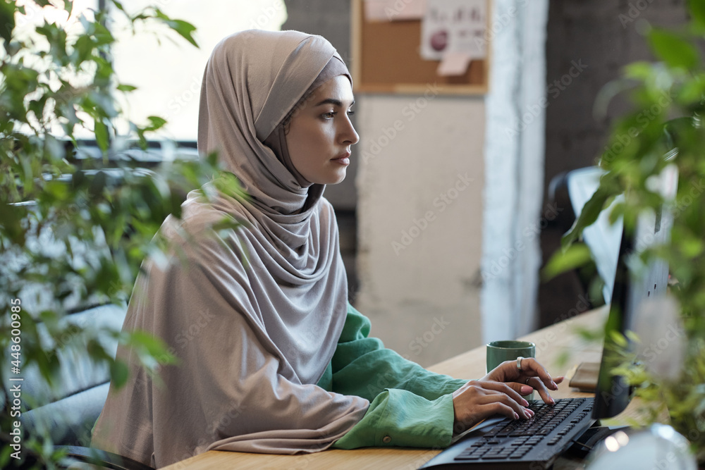 Poster serious muslim female in hijab looking at computer screen while networking