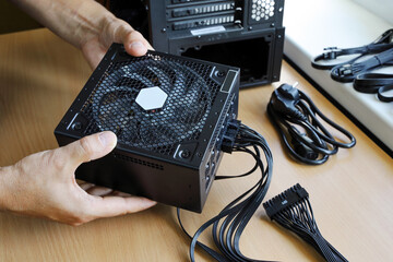 Hands hold power supply unit with connected wires to install it in computer case - obrazy, fototapety, plakaty