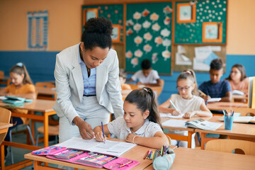 Black elementary school teacher assisting her student on class in classroom.