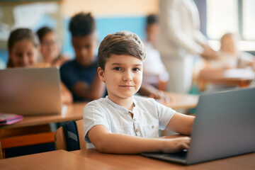 Elementary student using laptop on class at school and looking at camera.