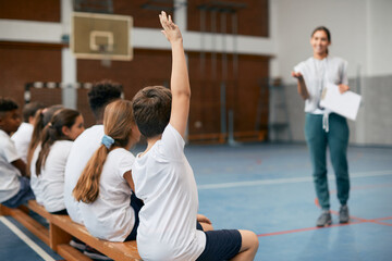 Rear view of schoolboy raising hand to ask question during physical education class.