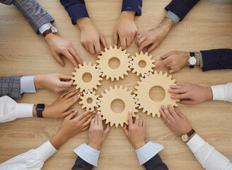 Team of business people join gearwheels. High angle, overhead view of circle of hands holding cogs on office table. Metaphor for good effective business system, cooperation, teamwork and efficiency - obrazy, fototapety, plakaty