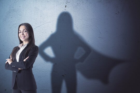 Attractive young european businesswoman with folded arms standing on concrete wall background with superhero cape shadow. Success and leadership concept.