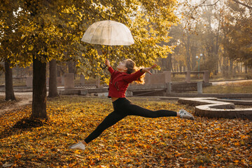 Hello fall, autumn season, fall mood, positive emotions. Teenager girl jumping with transparent umbrella and enjoying life in autumn sunny park