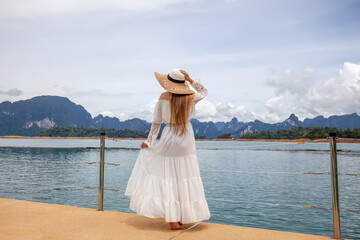 Back view of Happy Travel Girl with Hat Fun on Wooden Pier with Lake, Rainforest Jungle and Mountains on Background. Female Tourist Enjoy Vacation Cheow Lan Lake in Khao Sok National Park in Thailand