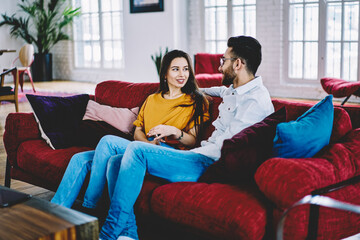 Carefree marriage in casual wear talking about flat design resting at cozy couch,Middle Eastern boyfriend discussing relationship with Caucasian girlfriend spending weekend in comfortable apartment