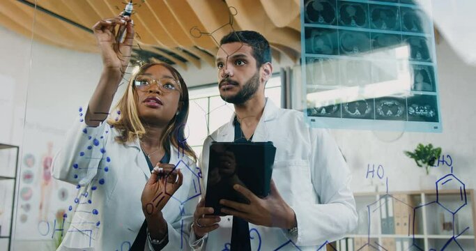 Attractive cvalified wise two diverse chemists in white gawns writing structural formula of chemical elements on glass wall in medical lab during joint work over research of drugs by genetic diseases