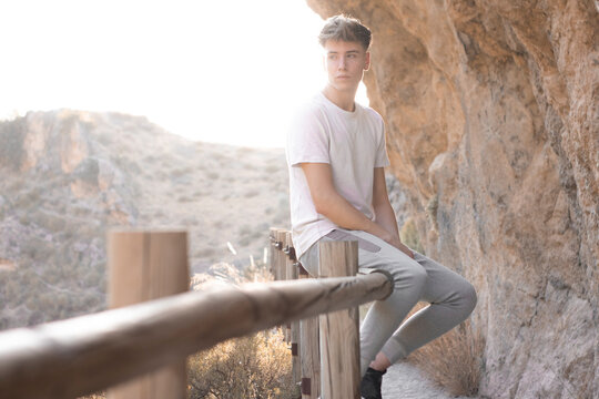 Hombre joven rubio pensativo sentado haciendo senderismo junto a las monta&ntilde;as en la puesta de sol