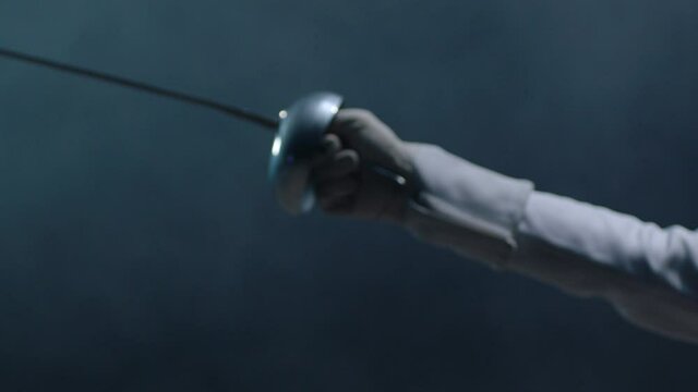 Professional fencer in protective uniform practicing maneuvers with sword alone in dark studio . Fencing demonstration on black smoked background . Shot on Arri Alexa cinema camera in slow motion .