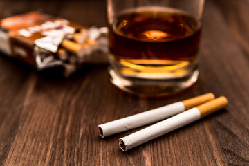 Glass of whiskey and two cigarettes with a pack of cigarettes on a wooden table. Focus on the cigarettes, shallow depth of field