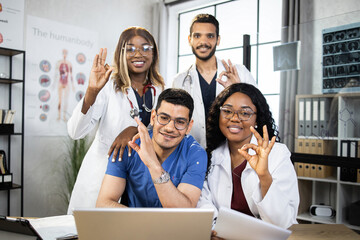 Positive male and female doctors in medical clothes posing at table with modern gadgets, smiling and showing okay gesture on camera. Multiracial specialists on consilium. Approval concept.