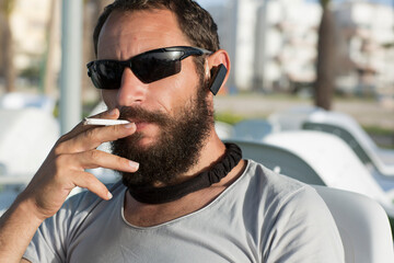 brutal Russian Caucasian beautiful white cool guy smoking wearing sunglasses and grey t-shirt in a cafe. American man with a cigarette in his hand. handsome Jewish man man mustache and beard, close up