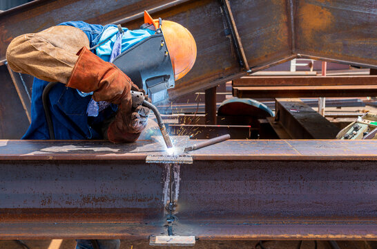 Welder Is Welding Add Joint H-beam For Steel Structure Work With Process Flux Cored Arc Welding(FCAW) And Dressed Properly With Personal Protective Equipment(PPE) For Safety, At Industrial Factory.