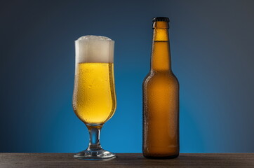 Unopened cold beer bottle and full beer glass on a wooden table blue background