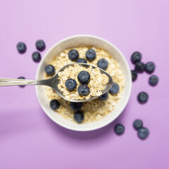 Creative concept still life health diet food photo of bowl dish with healthy breakfast oatmeal corn flackes fruits berries blueberry banana  yoghurt on purple background.