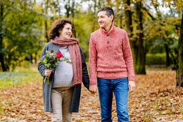 Husband with his pregnant wife holding by hands while walking in autumn park.