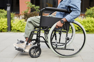 Unrecognizable black invalid in casual outfit holding wheelchairs handrails and pushing towards wheelchair outdoors