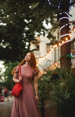 Young beautiful red-haired long-haired woman in a summer pink dress walks through the streets of a summer city.