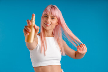 Portrait of young woman with pink hair smiling and showing bottle of moisturizing spray to camera...