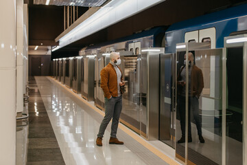 A man in a face mask is holding a smartphone while waiting for a subway train.