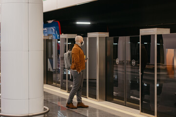 A man in a face mask is holding a smartphone while waiting for a subway train.