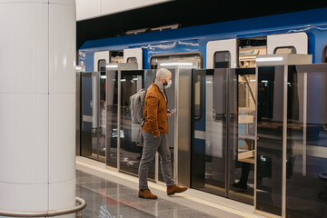 A man in a medical face mask is holding a smartphone while entering a subway car