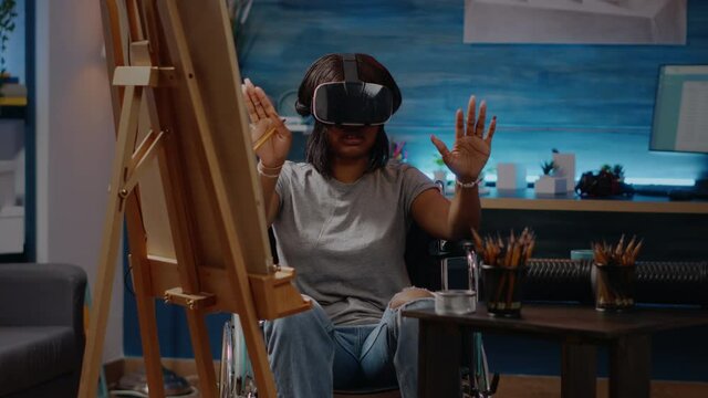 Black invalid artist woman using vr glasses for art project in workshop room at home. African american person with handicap sitting in wheelchair while having headset for masterpiece