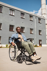 Fototapeta na wymiar Serious depressed young African-American man in eyeglasses sitting in wheelchair against gray building and looking into distance