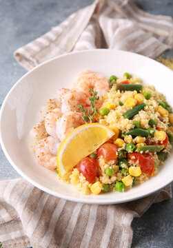 Seafood dish. Shrimp with couscous and vegetables: asparagus beans, corn, tomatoes, green peas and lemon in a white plate on grey table. Background image, copy space