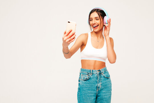 Beautiful smiling woman dressed in jeans clothes. Sexy carefree model listening music in wireless headphones. Adorable and positive female posing on grey background in studio.Taking selfie photos