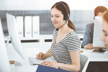 Obraz premium Casual dressed young woman using headset and computer while talking with customers online. Group of operators at work in sunny office. Call center