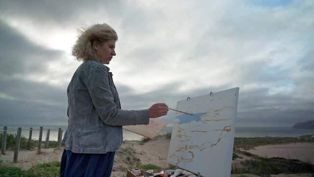 Middle-aged European woman artist paints a picture with paints overlooking the sea. Blue skies. Close-up.