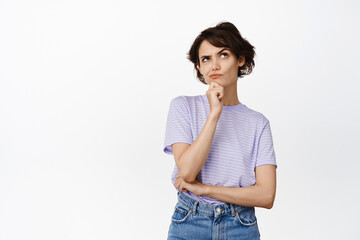 Image of pretty thoughtful girl ponder, looking up serious and thinking, makes decision, touching chin, choosing, standing over white background. Copy space