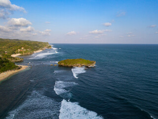 Drone photo of the beach with overgrown by lush green trees with waves rolling towards the shoreline and coral islands off the coast