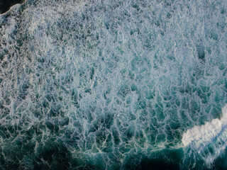 Drone photo of The dramatic lapping of the waves in the ocean produces white foam. The sea water of the pond is blue and contrasts with the color of the foam